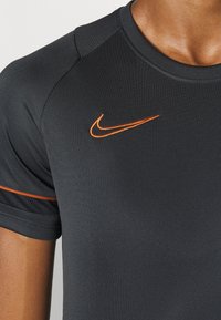 Närbild på en person som bär en mörk träningsskjorta med en orange Nike swoosh-logotyp på bröstet och en orange rand på ärmen.