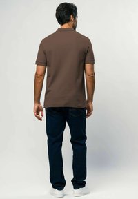 Tricou polo maro cu guler, mâneci scurte, asortat cu blugi de culoare albastru închis și adidași albi, văzut din spate.