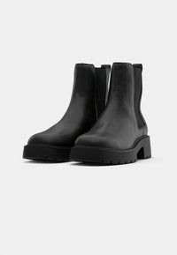 Botas de tornozelo em couro preto com sola de borracha espessa, painéis laterais elásticos e uma textura suave, apresentando um design minimalista.