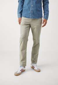 Pantaloni grigi chiari con una texture liscia, abbinati a sneaker bianche con righe rosse e suola in gomma. Indossa anche una camicia in denim blu.