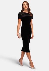 Abito in velluto nero con un cappotto off-the-shoulder in paillettes, silhouette aderente, taglio al ginocchio, abbinato a tacchi argentati e una clutch.
