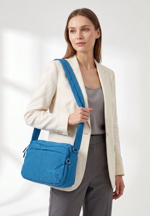 Femme en blazer beige et pantalon gris tenant un sac à bandoulière bleu avec logo GMV sur fond blanc uni.