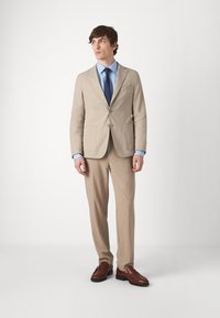 Completo beige chiaro con giacca a un bottone, pantaloni abbinati e scarpe in pelle marrone. Una camicia azzurro chiaro e una cravatta blu navy completano l'outfit.