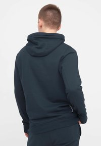 Haut à capuche bleu marine foncé avec une capuche à cordon de serrage, texture lisse, poignets ajustés et un accent logo discret près de l'ourlet. Vue arrière montrée.