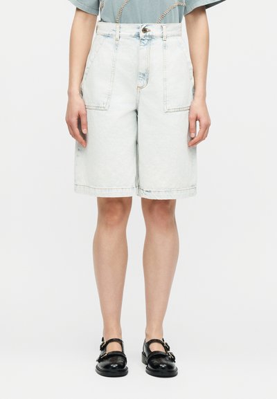 Άτομο που φοράει ανοιχτόχρωμα τζιν σορτς με ψηλή μέση και μήκος μέχρι το γόνατο, με μεγάλες μπροστινές τσέπες και μαύρα παπούτσια με αγκράφα, στέκεται σε λευκό φόντο.
