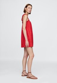 Piros ujjatlan ruha texturált anyagból, fodros pántokkal, hímzett részletekkel és hullámos szegéllyel. Barna szandállal párosítva.