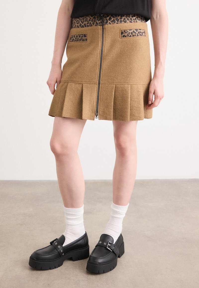 Femme portant une jupe plissée marron avec une ceinture et des poches à imprimé léopard, des mocassins noirs épais, des chaussettes blanches, debout sur un sol gris.