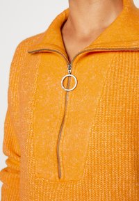 Close-up of a person wearing a bright orange knit sweater with a partially zipped silver zipper featuring a round pull tab.