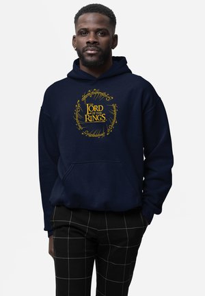 Mand iført marineblå hættetrøje med gul tekst "The Lord of the Rings" og ring-skriftdesign, kombineret med sorte bukser med hvide gitterlinjer.