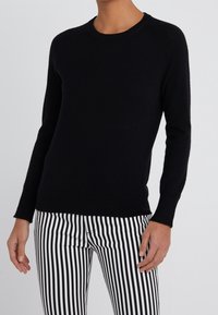 Schwarzer Strickpullover mit rundem Halsausschnitt und langen Raglanärmeln. Getragen mit schwarz-weißen gestreiften Hosen, die eine glatte Textur und Passform zeigen.
