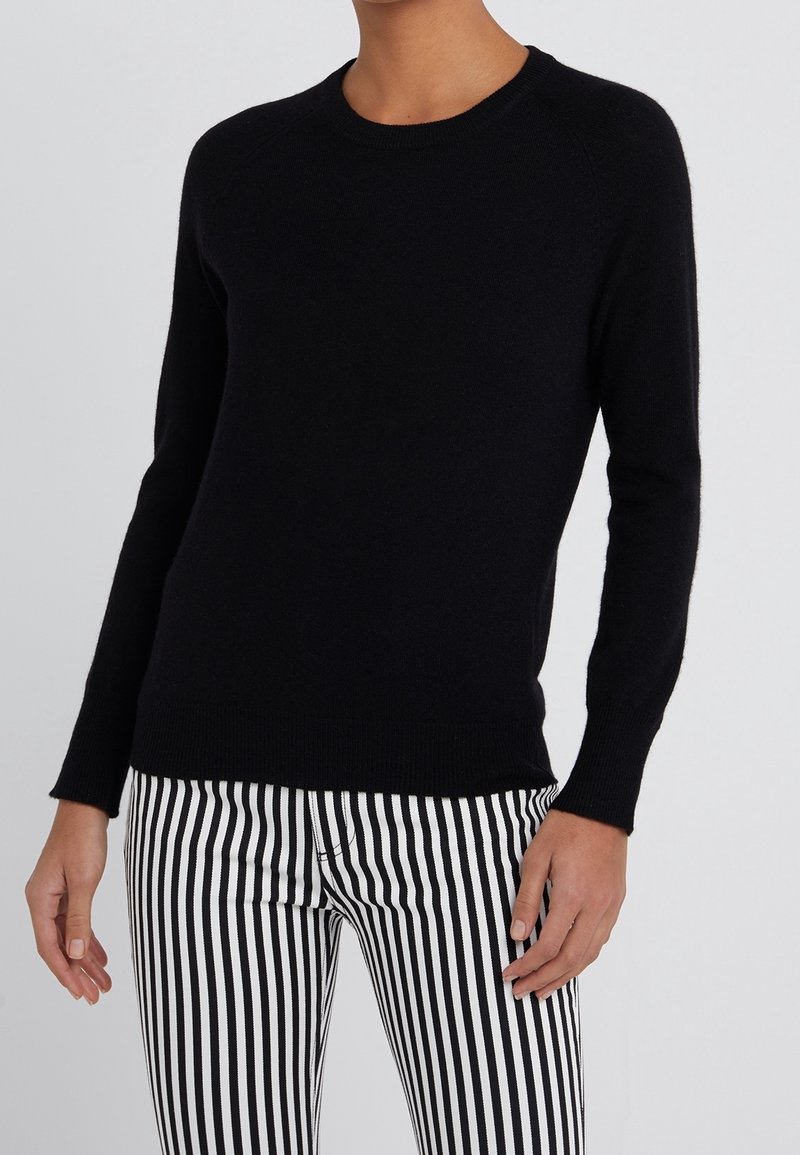 Schwarzer Strickpullover mit rundem Halsausschnitt und langen Raglanärmeln. Getragen mit schwarz-weißen gestreiften Hosen, die eine glatte Textur und Passform zeigen.