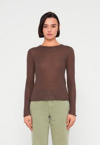 Καφέ μπλούζα μακρυμάνικη σε εφαρμοστή γραμμή με ριμπ ύφασμα και κυματιστό τελείωμα; συνδυασμένη με ανοιχτό πράσινο τζιν, σε απλό φόντο.