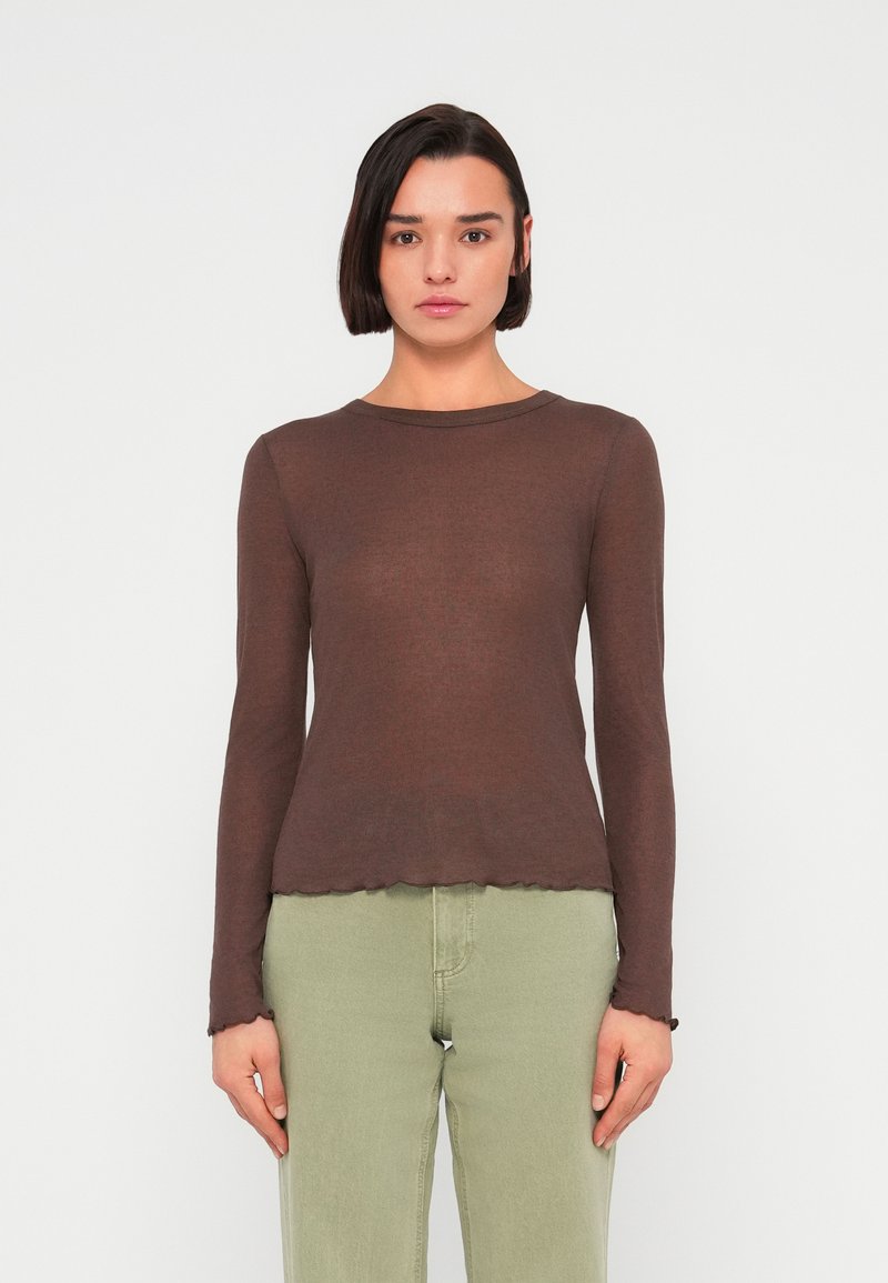 Καφέ μπλούζα μακρυμάνικη σε εφαρμοστή γραμμή με ριμπ ύφασμα και κυματιστό τελείωμα; συνδυασμένη με ανοιχτό πράσινο τζιν, σε απλό φόντο.