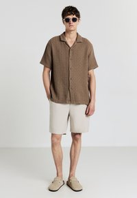 Braun gemustertes Kurzarmhemd, beige gestreifte Shorts und hellbeige Slippers. Runde Sonnenbrillen vervollständigen das Outfit.