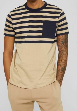 Hombre con camiseta de manga corta a rayas beige y azul marino con bolsillo azul marino y pantalones beige, manos en los bolsillos, mostrando el brazo derecho tatuado.