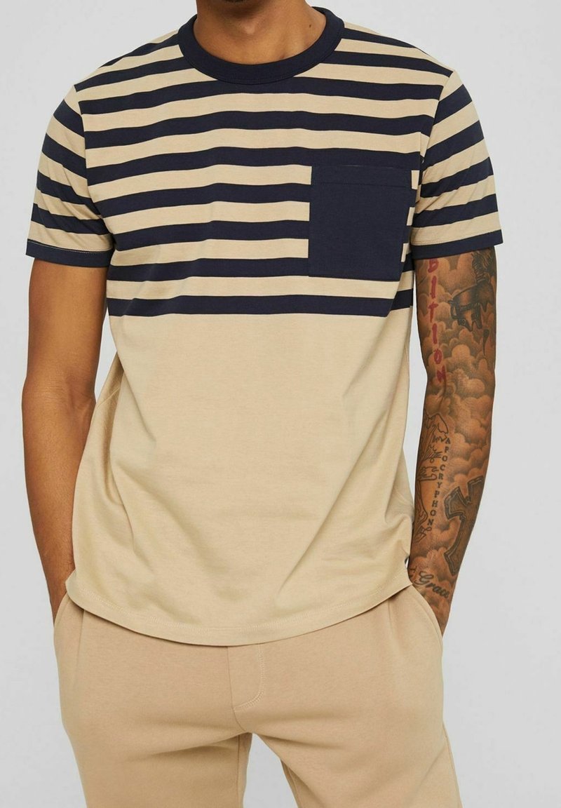 Homme portant un t-shirt à manches courtes beige et bleu marine rayé avec une poche bleu marine et un pantalon beige, mains dans les poches, montrant un bras droit tatoué.