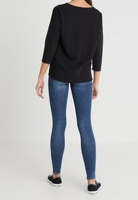 Schwarzes Baumwolloberteil mit Dreiviertelärmeln, kombiniert mit eng anliegender blauer Jeans und schwarzen Sneakers mit weißen Akzenten. Rückansicht dargestellt.