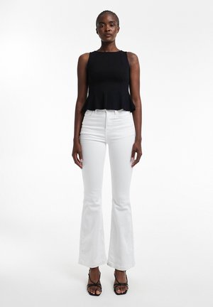 Femeie în picioare purtând un top negru fără mâneci cu peplum, pantaloni albi evazați cu talie înaltă și sandale negre cu toc și barete.