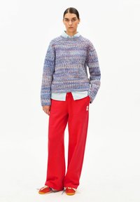 Gebreide trui in blauw- en paarstinten met een gestructureerd patroon, gecombineerd met losse rode broek met een wit logo, en oranje sneakers.