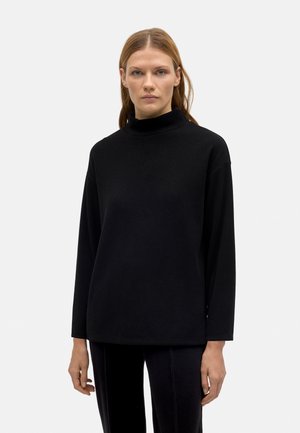 Top de cuello alto negro con mangas largas, que presenta un corte relajado y una textura suave, sobre un fondo claro liso.