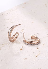 Roségoldene Creolen mit einem gedrehten Design, versehen mit kleinen klaren Kristallen entlang einer Kante, die ein poliertes Metallfinish zeigen.