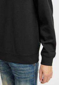 Zwarte sweatshirt met een ronde halslijn, ribgebreide manchetten en onderrand; gecombineerd met blauwe versleten jeans. Eenvoudige, gladde stofstructuur.