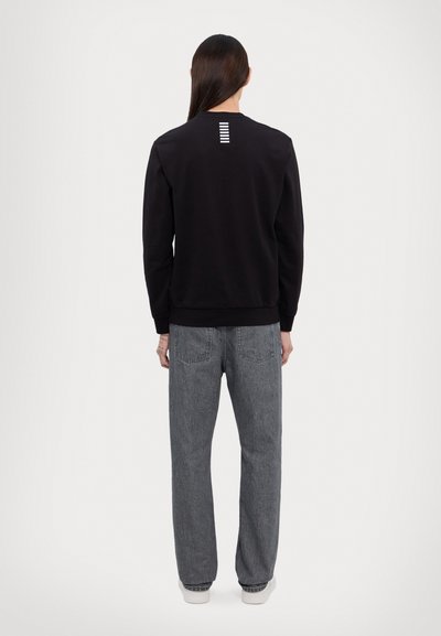 Sweatshirt noir avec des poignets et un ourlet côtelés, présentant des rayures blanches verticales au dos. Assortie avec un jean en denim gris taille haute.