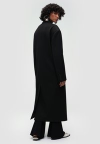 Cappotto lungo nero con taglio dritto, caratterizzato da uno spacco centrale sul retro e dettaglio con bottoni sulle maniche. Tessuto liscio e sartoriale.