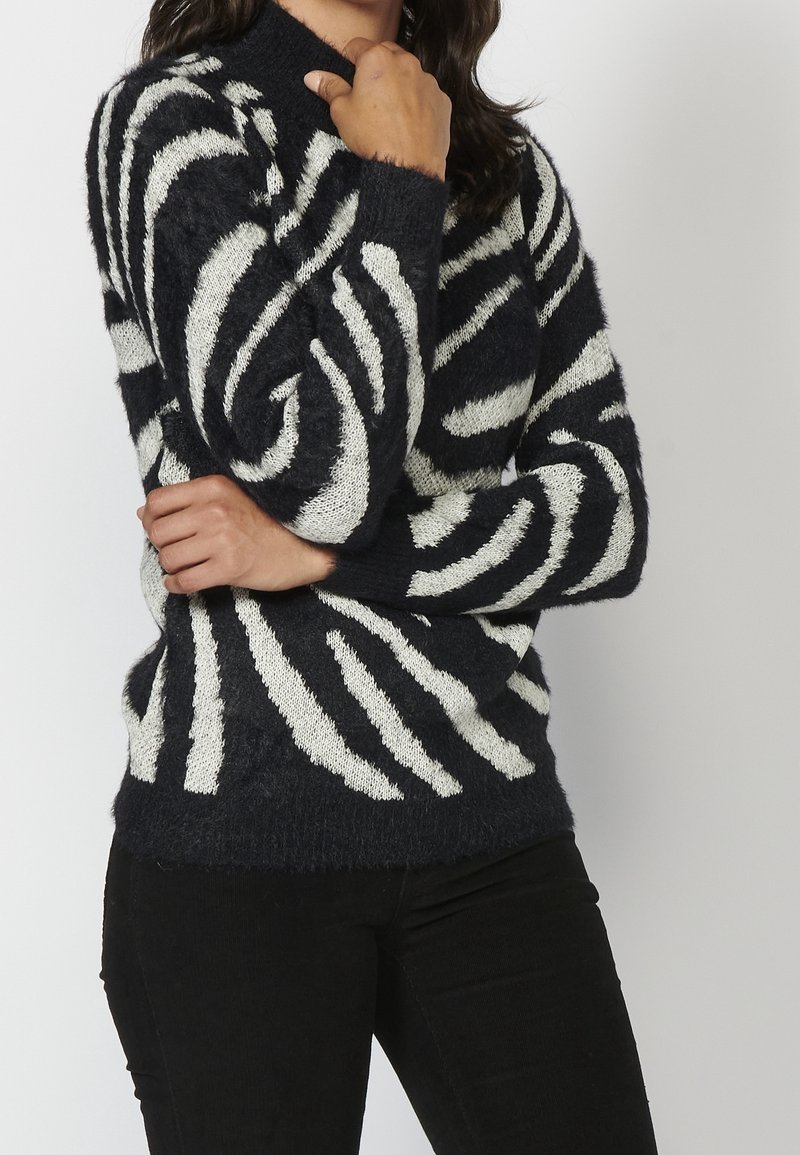 Personne portant un pull douillet à rayures zébrées noires et blanches à manches longues et un pantalon noir, posant les bras croisés.