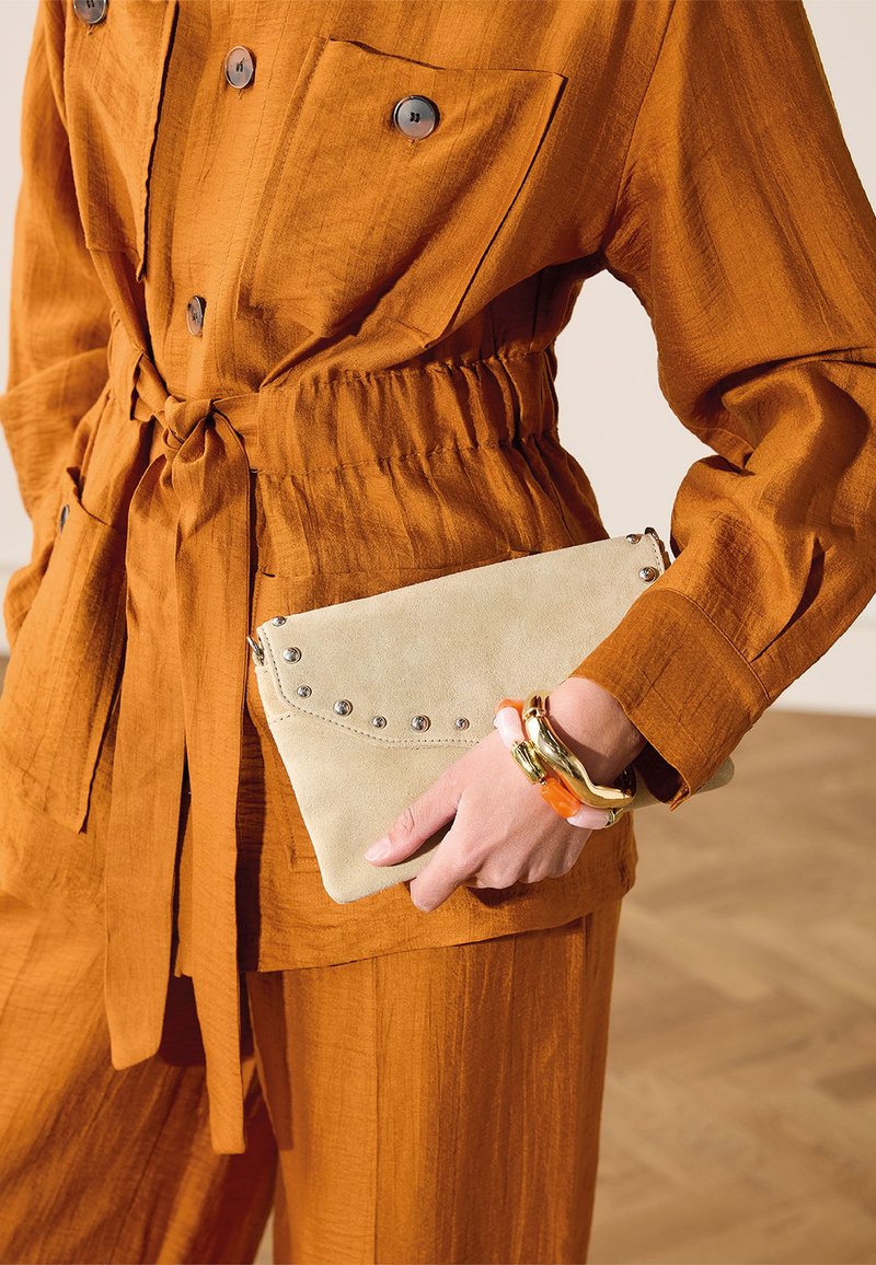 Persoon draagt een roestkleurige outfit met knopen en taillekoord, houdt een beige gestikte clutch vast en draagt kleurrijke dikke armbanden om de pols.