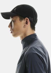 Sort atletisk cap med perforeret design, flad skygge og et logo foran. Båret med en mørk, let jakke. Profilbillede fra siden.