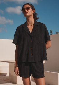Black short-sleeve button-up shirt with a pocket and textured pattern, paired with matching shorts. Accessories include brown sunglasses.