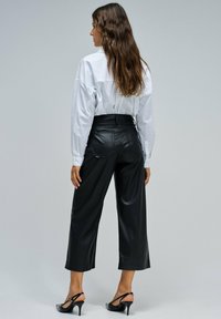Mujer con cabello largo y ondulado, vestida con una camiseta blanca de manga larga, pantalones de pierna ancha de tiro alto negros y tacones tipo slingback negros, de pie de lado.