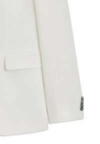 Manga de blazer blanca con cuatro botones oscuros en el puño y un bolsillo con solapa en el costado, resaltando los detalles y la textura de la prenda.