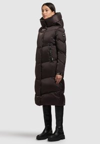 Langer, dunkelbrauner Steppmantel mit großem Kragen, Reißverschlusstasche, strukturiertem Steppmuster und mattem Finish; kombiniert mit schwarzen kniehohen Stiefeln.