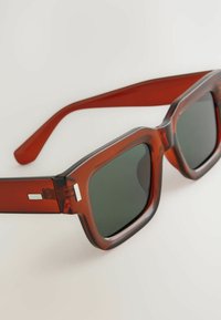 Lunettes de soleil rectangulaires avec une monture transparente rouge foncé, des verres vert foncé, des accents en métal et des branches épaisses. Texture lisse sur l'ensemble.