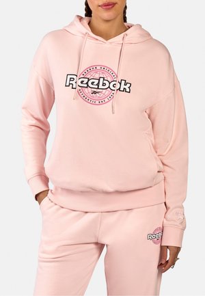 Frau trägt hellrosa Reebok-Hoodie und passende Jogginghose mit Logodrucken, Hände in den Taschen, steht vor schlichtem Hintergrund.