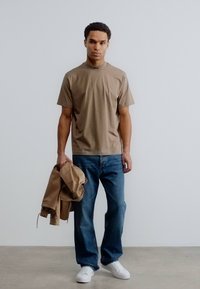 Κοντομάνικο μπλουζάκι σε ταμπά χρώμα, μπλε τζιν παντελόνι και λευκά αθλητικά παπούτσια. Το μοντέλο κρατάει ένα ανοιχτό καφέ μπουφάν, στέκεται σε γκρι δάπεδο μπροστά από λευκό φόντο.