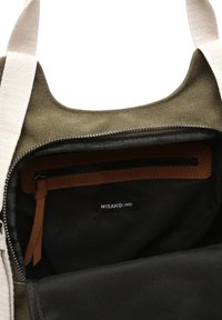 Interiér olivově zelené plátěné tašky s černou podšívkou, obsahující malou hnědou kapsu na zip a štítek s nápisem "MISAKO". Viditelné bavlněné popruhy.
