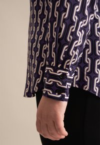 Camicia a maniche lunghe con un motivo a catena viola e bianco, con bottoni neri sui polsini e una texture del tessuto liscia.