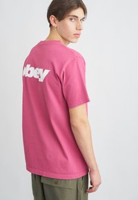 Krátký rukáv růžové bavlněné tričko s bílým logem "obey" na zádech, doplněné olivovými cargo kalhotami.