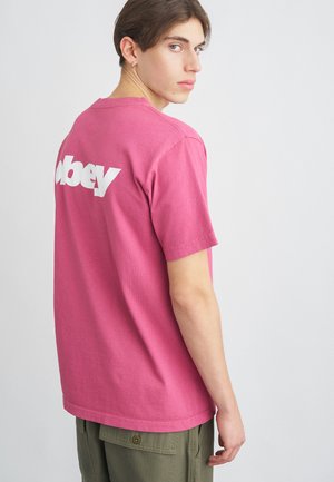 Rožinė medvilninė trumpomis rankovėmis marškinėliai su balta "obey" logotipu ant nugaros, derinami su alyvuogių žalumos krovinių kelnėmis.