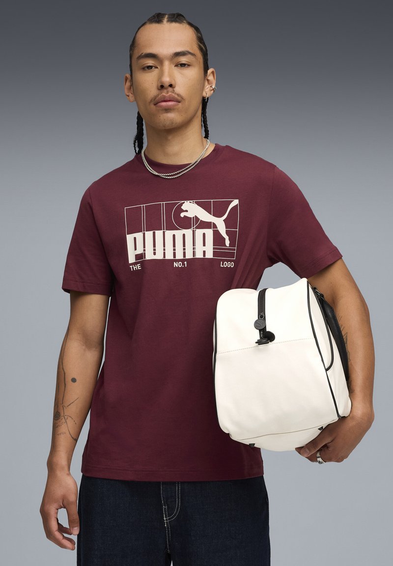 T-shirt bordeaux avec logo Puma blanc et texte ; sac blanc avec des détails noirs et fermeture éclair ; jeans foncés ; présenté sur un fond gris.