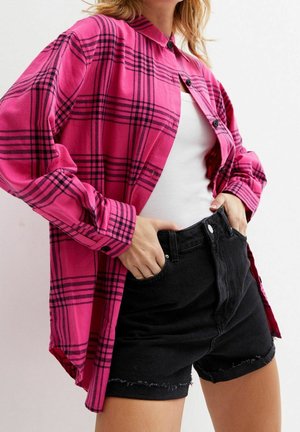 Vrouw draagt een felroze geruite blouse over een wit hemdje met zwarte hooggetailleerde denimshorts, handen in de voorzakken.