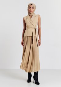 Abito plissettato beige con un corpetto aderente doppiopetto e cintura in vita; abbinato a stivali neri con tacco alto e a un hijab beige coordinato.