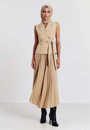 Abito plissettato beige con un corpetto aderente doppiopetto e cintura in vita; abbinato a stivali neri con tacco alto e a un hijab beige coordinato.