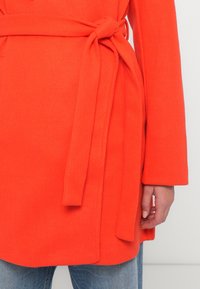 Close-up de uma pessoa vestindo um casaco laranja atado à cintura com um cinto, combinado com jeans azuis e mostrando uma mão relaxada.