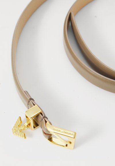 Ceinture en cuir beige avec une finition brillante et une boucle en métal doré ornée d'un motif d'aigle. La sangle est lisse avec une texture minimale.