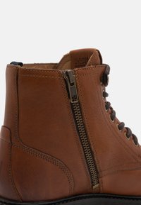 Brahna ādas zābaks ar sānu rāvējslēdzēju, brūnām auklām un teksturētu virsmu. Šuvju detaļas un melna zole.