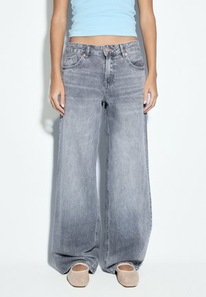 Person trägt ausgewaschene graue Jeans mit weitem Bein, hellblaues bauchfreies Oberteil und beige flache Schuhe vor schlichtem Hintergrund.